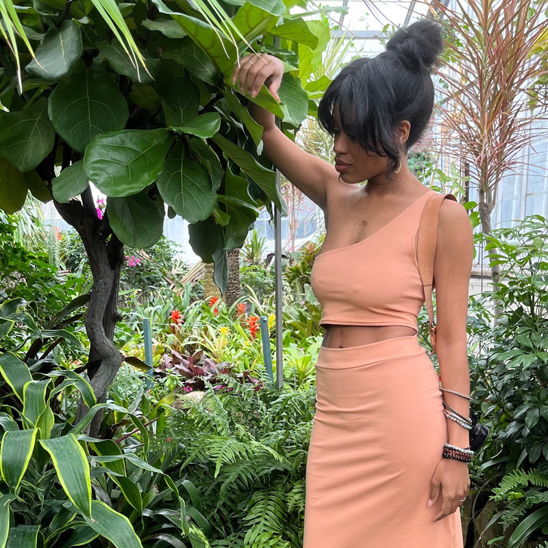 woman in tropical plant greenhouse wearing natural fiber asymetrical cropped tank top + skirt in peach colour