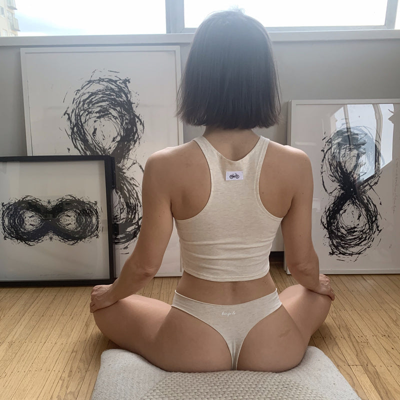 Cotton thong underwear + cropped bra top on woman meditating on cushion in front of artwork